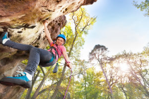 Bottom view of teenage rock climber exercising in the forest area against blue sky with copy-space